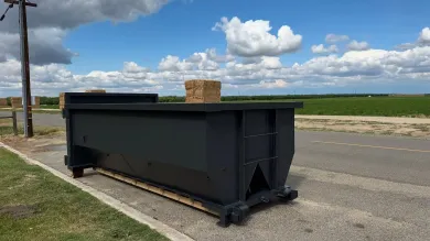 Black dumpster on a roadside, with hay bales in the background, under a blue sky.