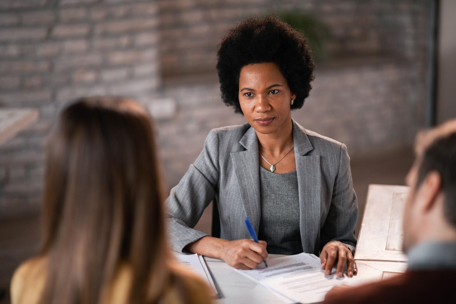 Meeting scene with a woman in a gray blazer taking notes across a table from two people