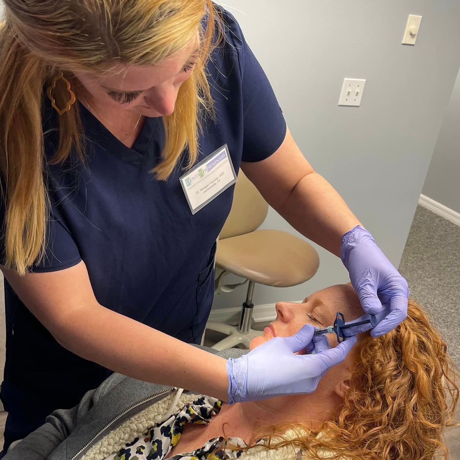 A person in blue scrubs injecting a woman's face. Both are indoors. The woman has red hair.