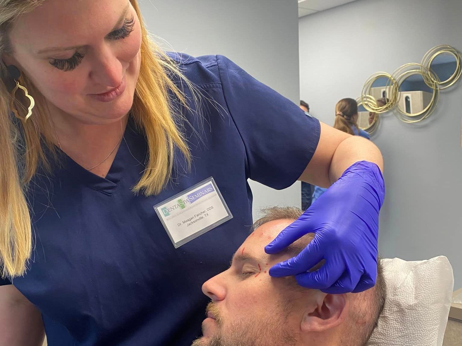 Medical professional examining a patient's forehead, wearing blue gloves and scrubs.