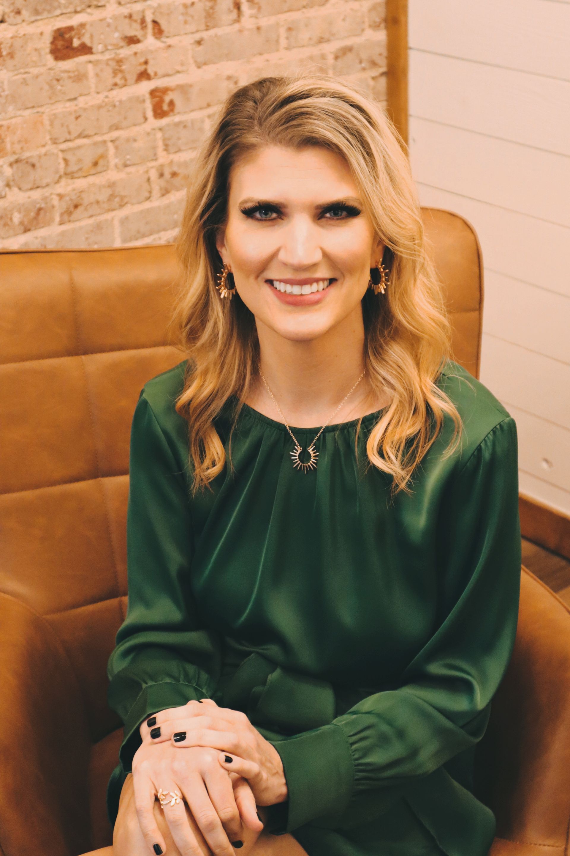 Woman in a green dress smiles, seated in a brown leather chair, against a brick and wood backdrop.