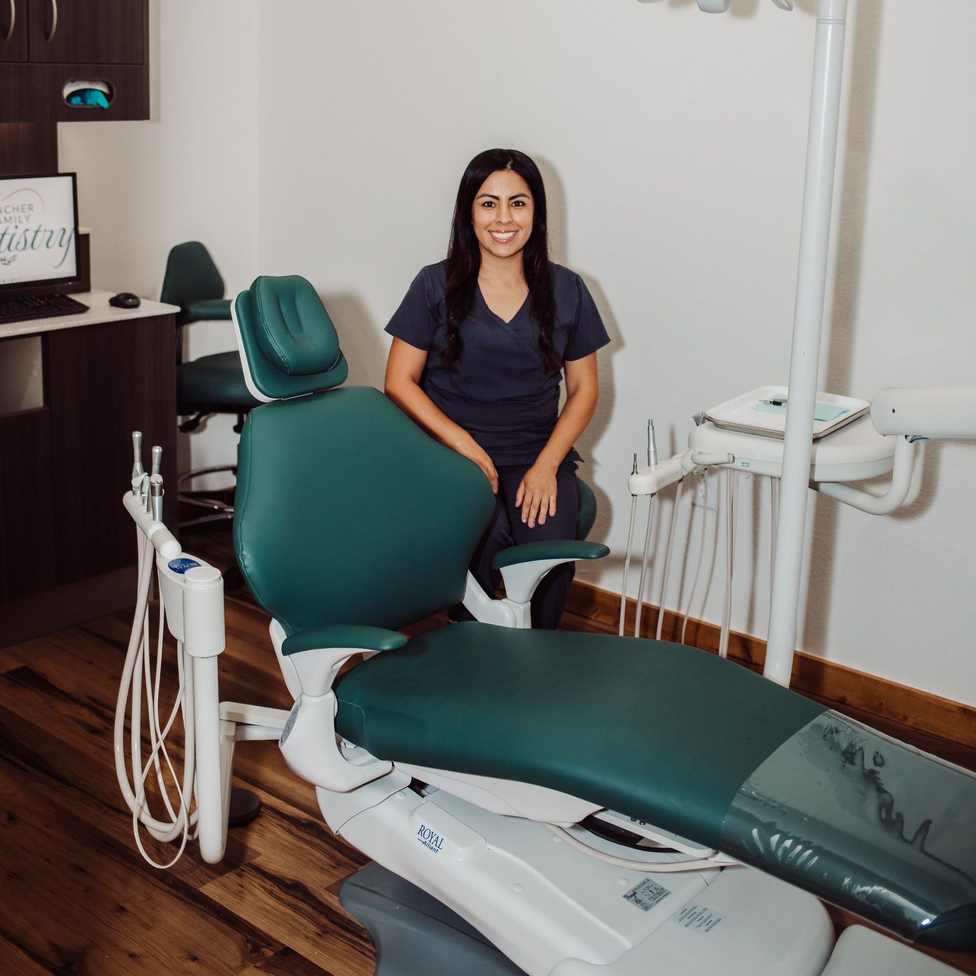 Dentist in blue scrubs kneels by a green dental chair in a bright office.