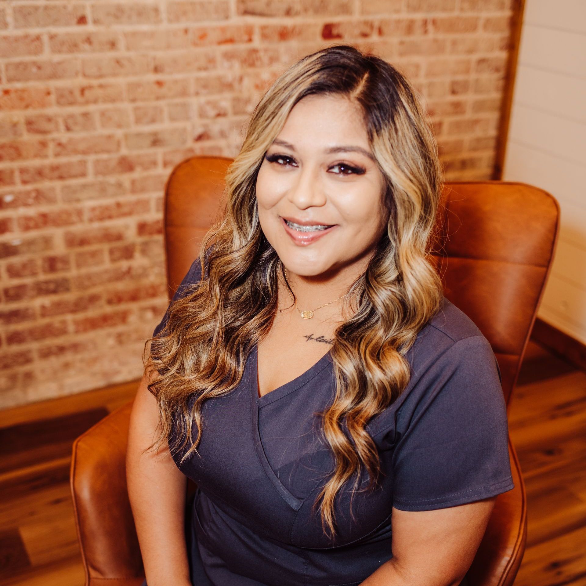 Woman in gray scrubs smiles, seated in a leather chair. Brick wall background.