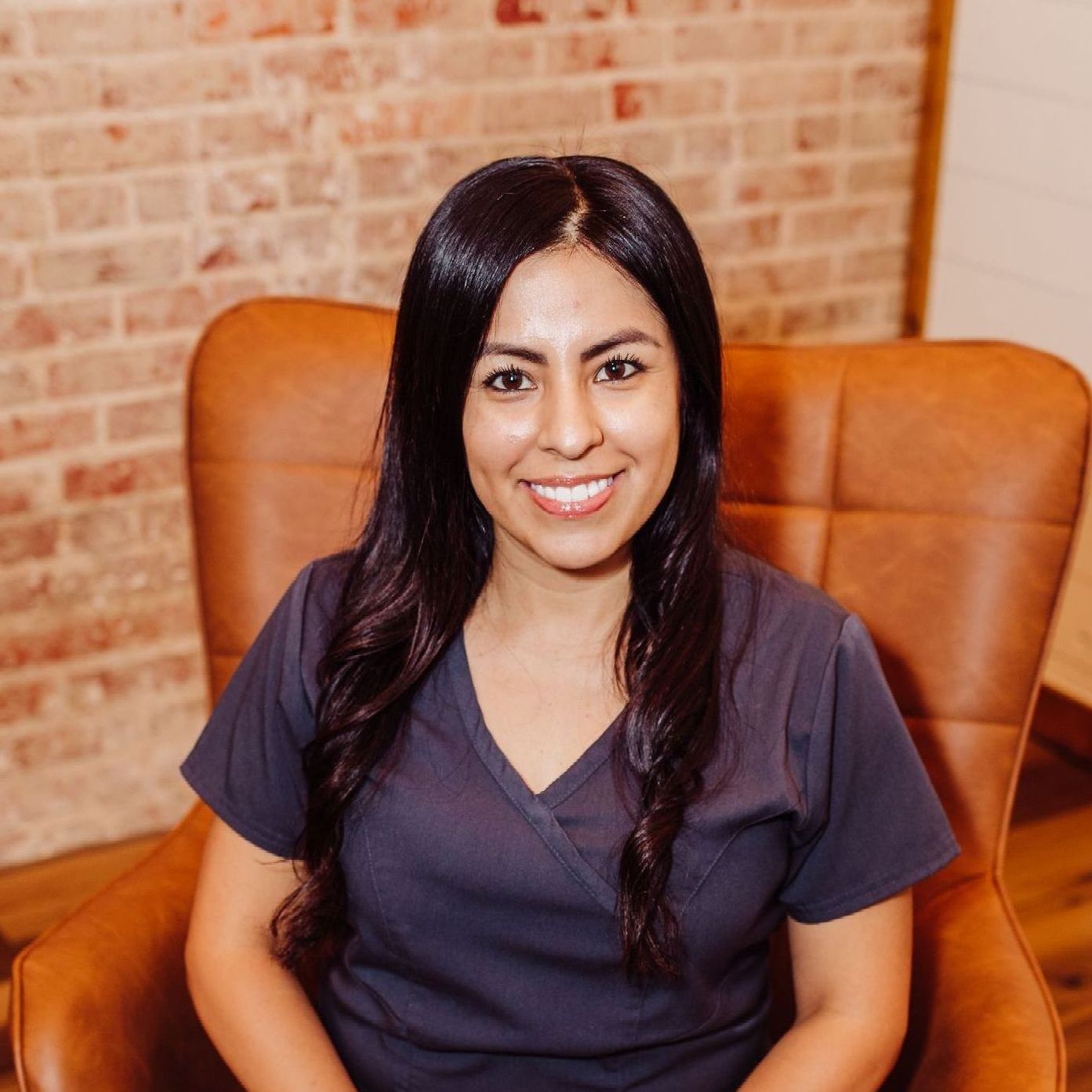 Woman in blue scrubs smiles, seated in a brown armchair, against a brick and wood wall.