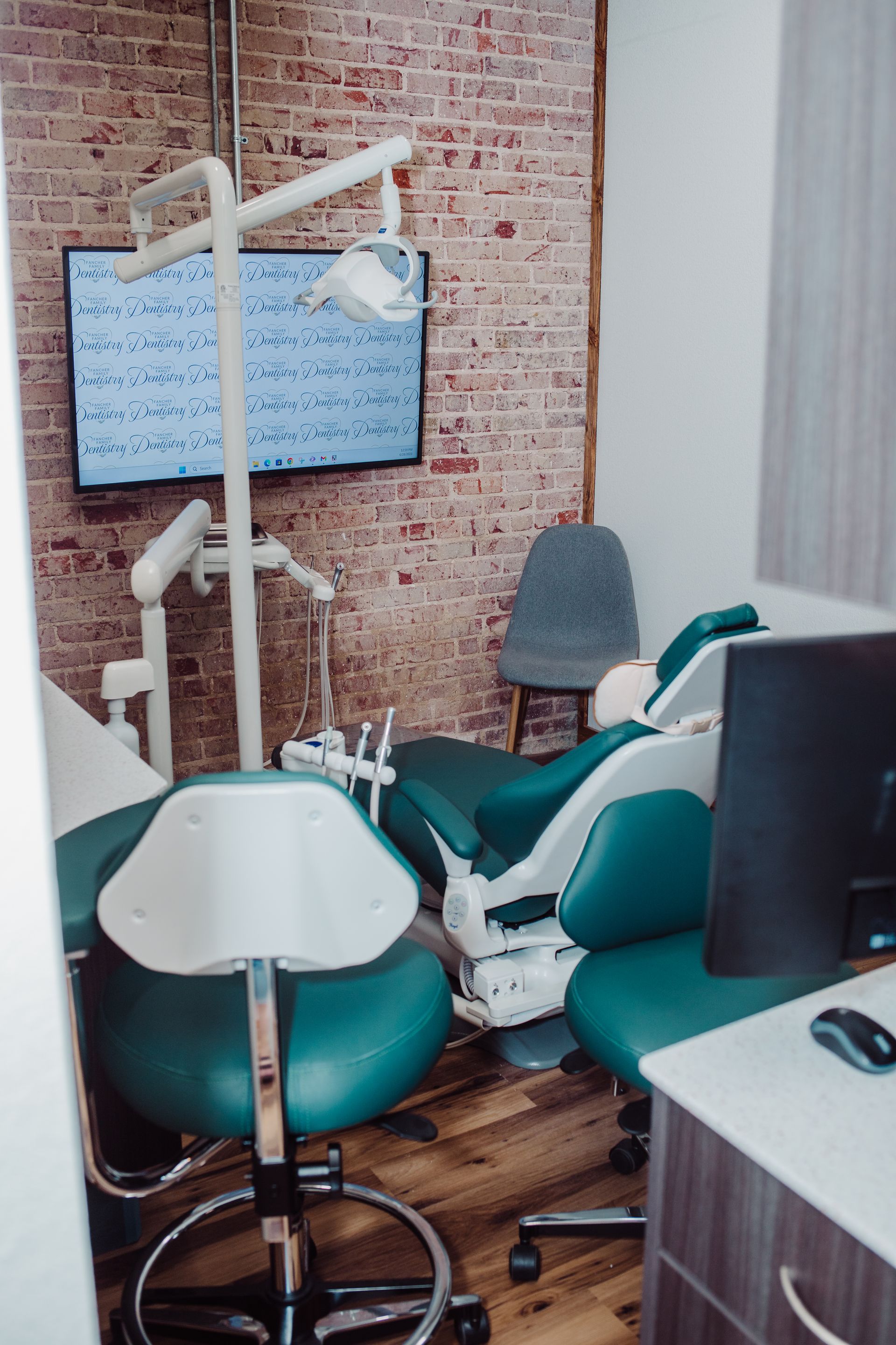 Dental exam room with teal chair, instruments, and a monitor on a brick wall.