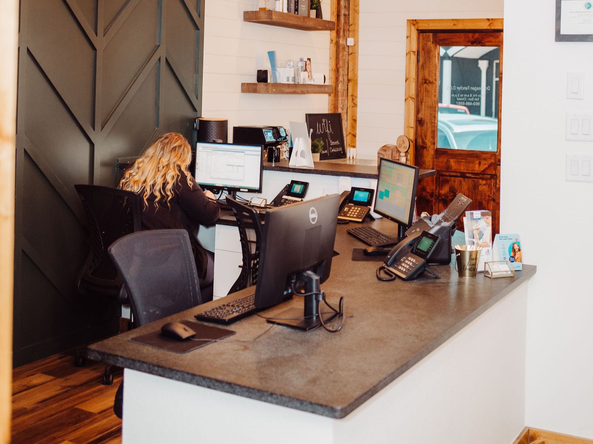 Reception desk with person working on a computer, phones, and items on the counter. Wooden accents, light walls.