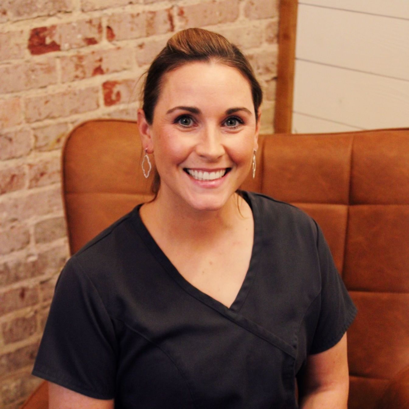 Woman smiling in a dark blue top, sitting in a brown chair, brick wall background.
