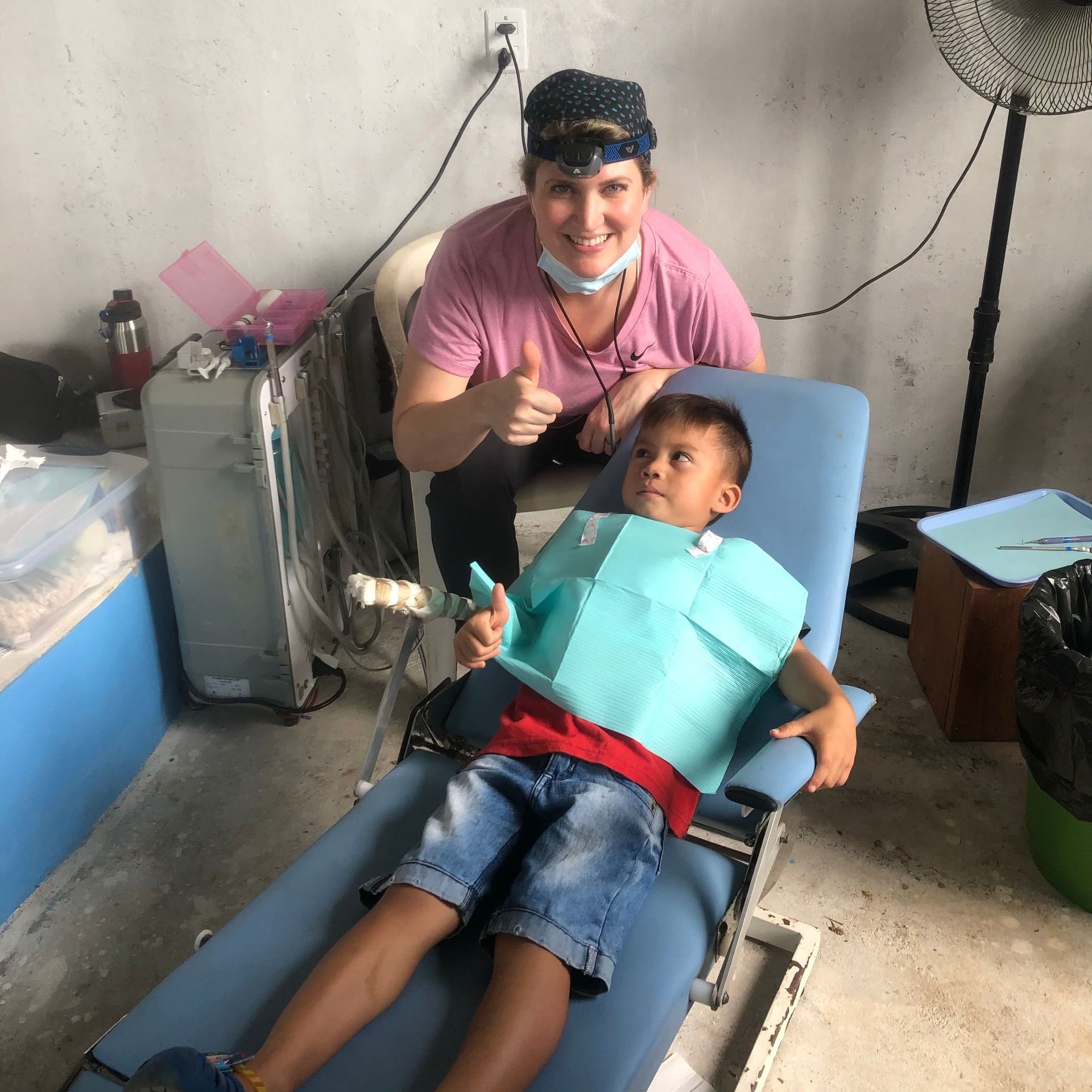 Woman in scrubs with child in dental chair; smiles, gives thumbs-up, medical setting.