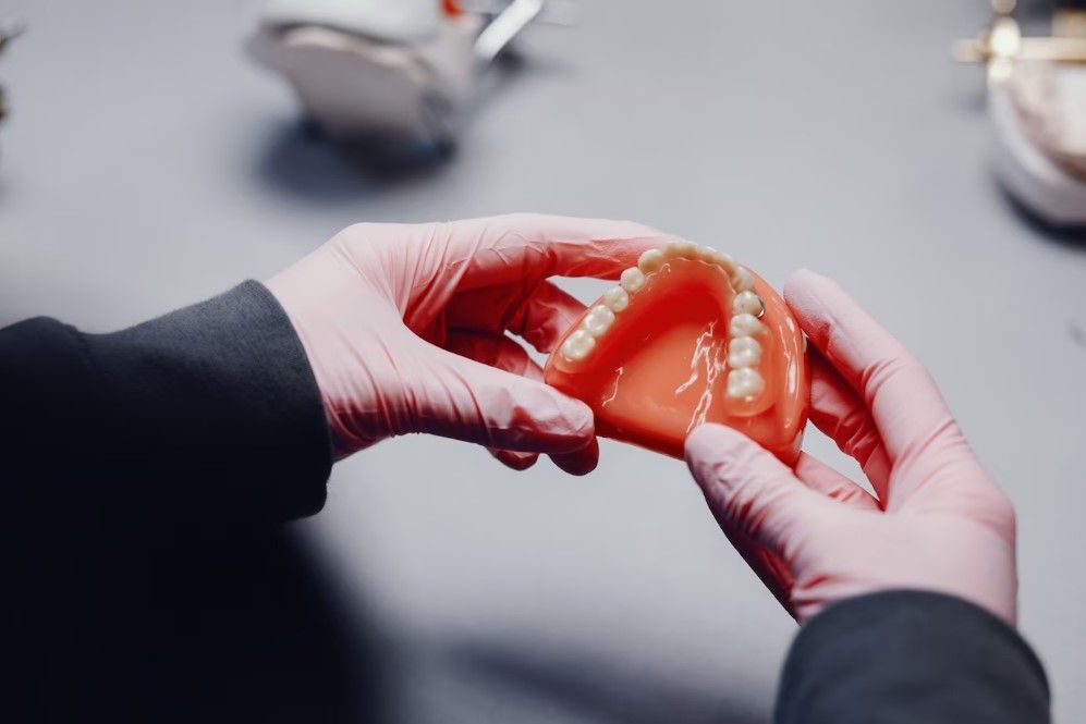 Hands in pink gloves holding a set of dentures, with other dental tools in the background.