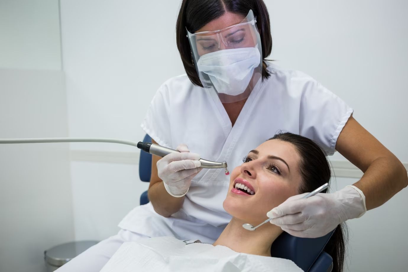 Dentist in mask, face shield, and gloves examines patient's teeth with dental tools in a bright, white-walled room.