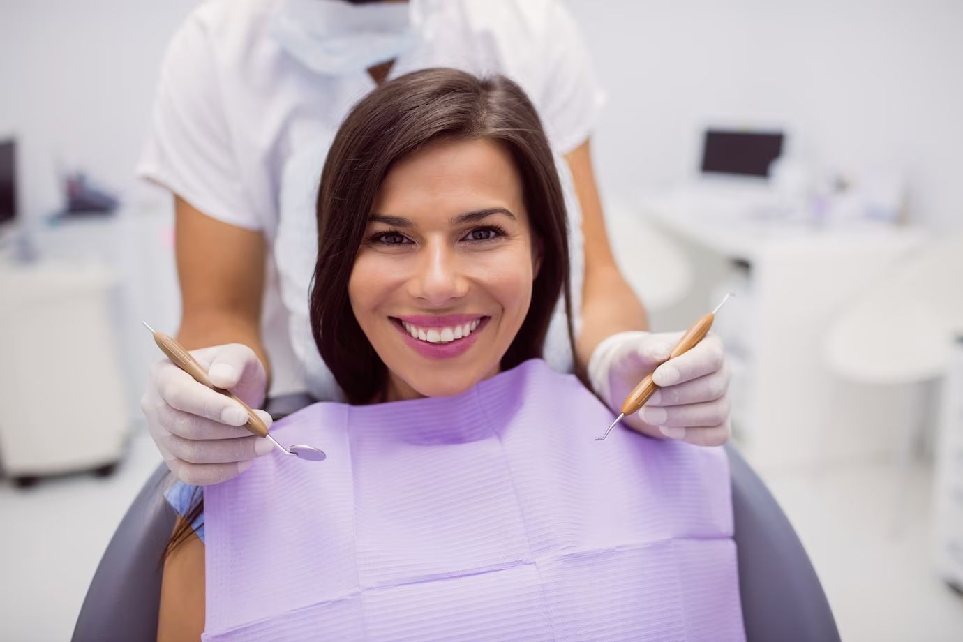 Woman smiling in a dentist's chair; dentist in white uniform holds dental tools.