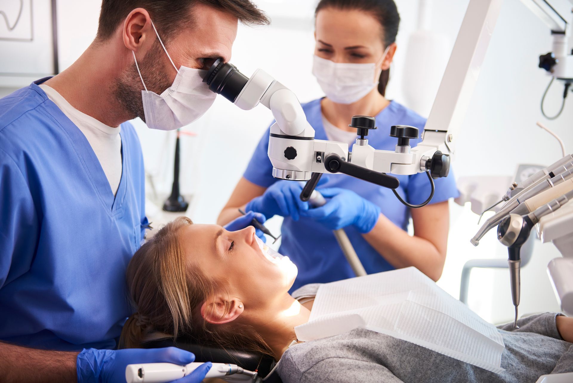 Dentist examining patient's teeth with microscope, dental assistant present.