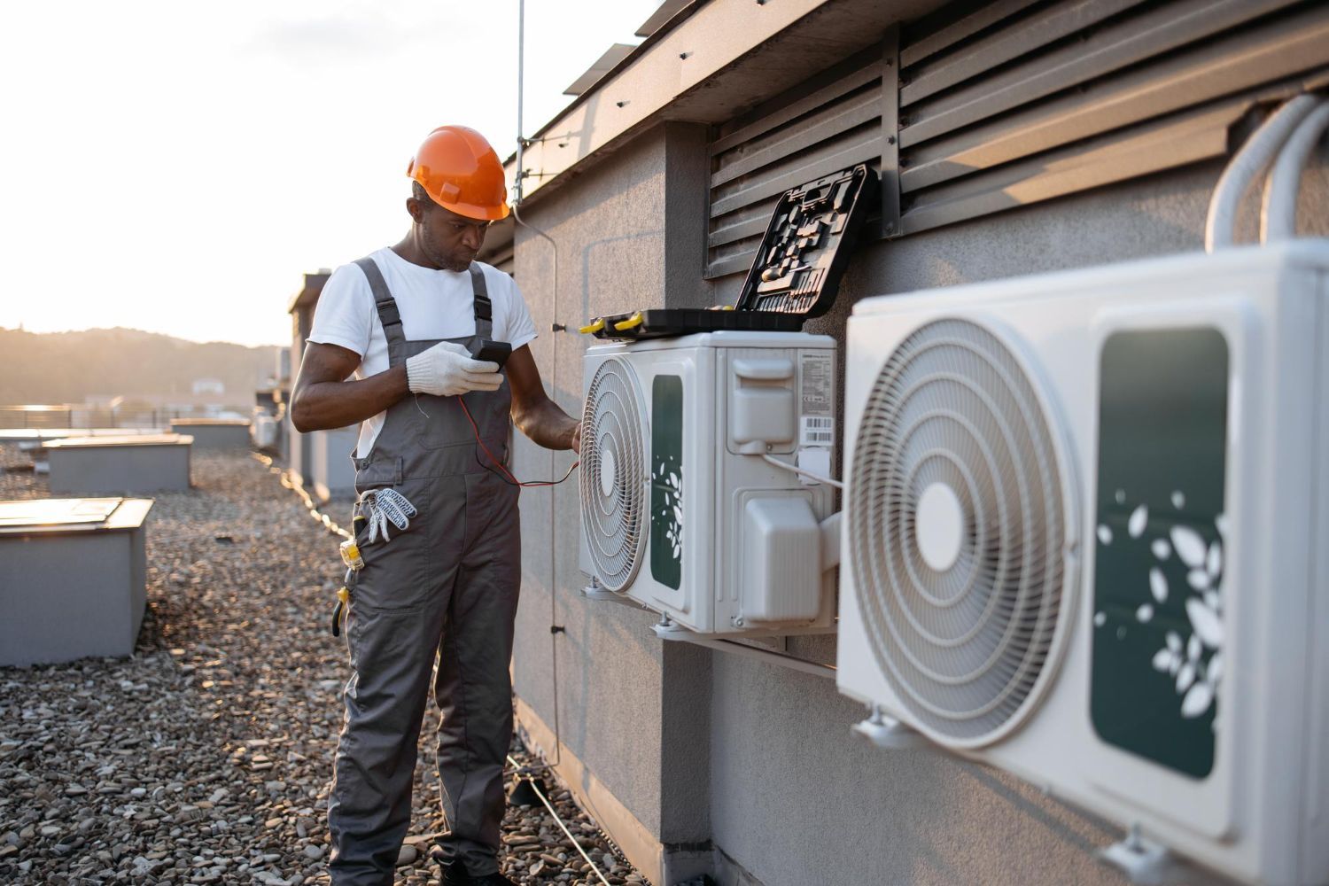 Technician inspecting outdoor HVAC units beside a building, holding a tablet and wearing a hard hat.
