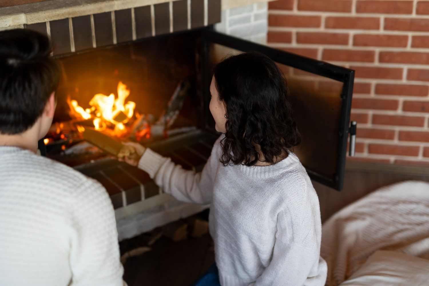 Two people sit by a lit fireplace, one tending the fire with a tool.