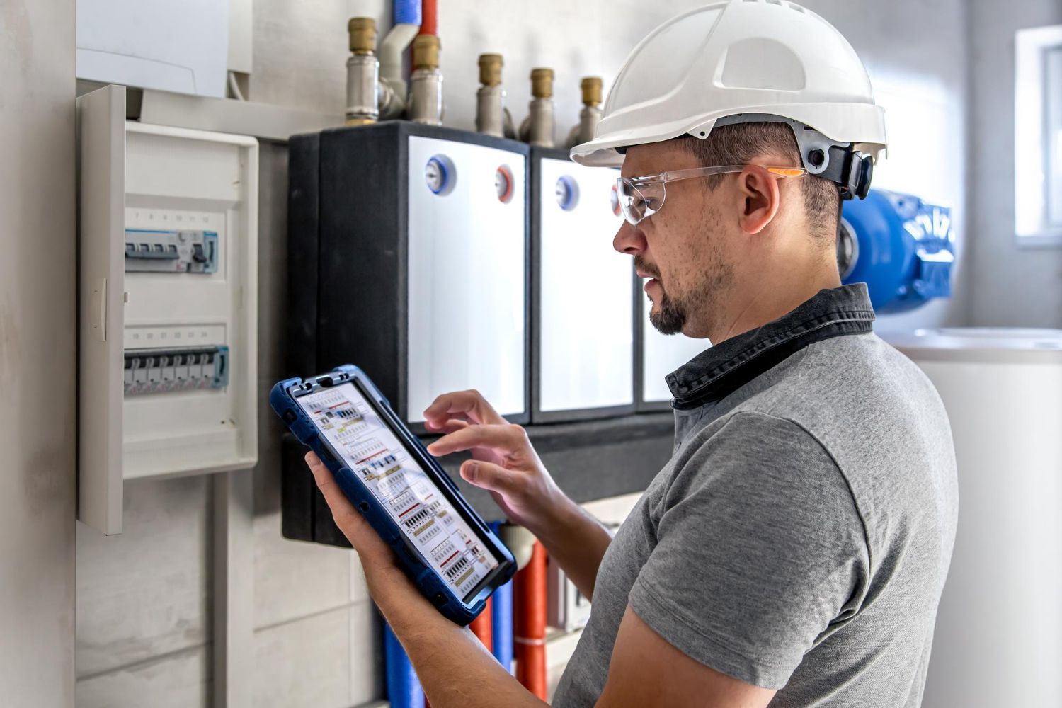 Technician in hard hat using a tablet to inspect electrical panels and pipes in a utility room