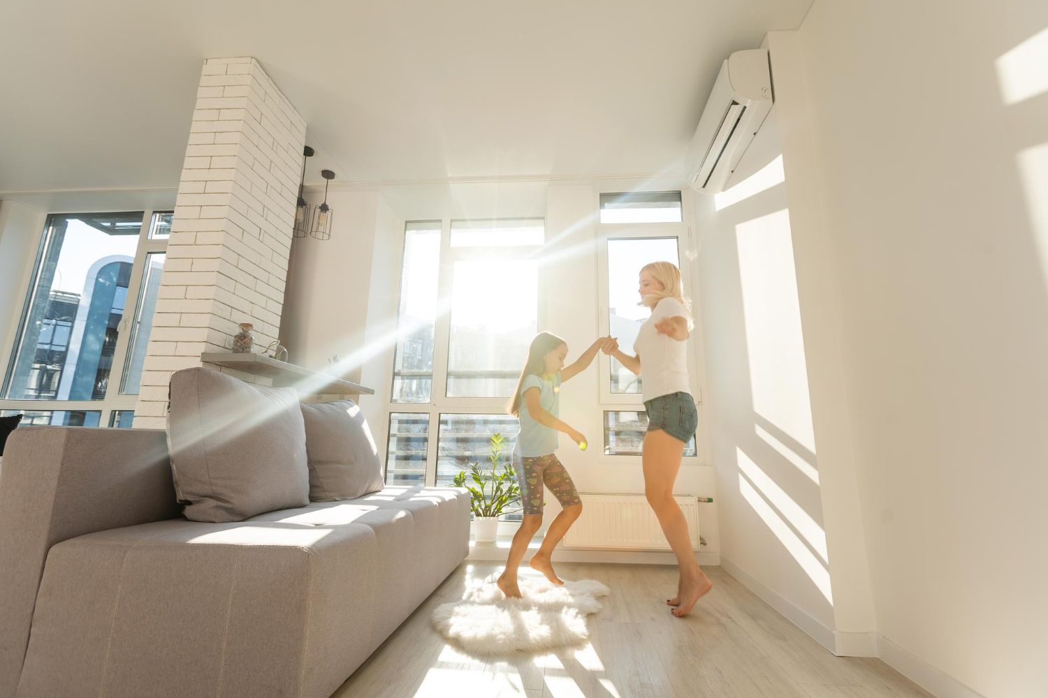 Two people dancing in a bright sunlit living room with large windows and a gray sofa
