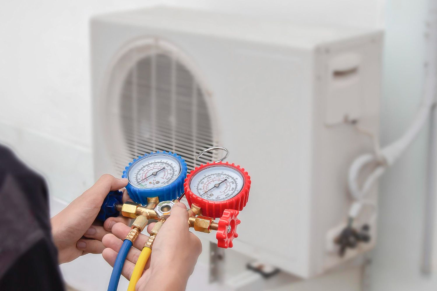 Hands holding blue and red gauges in front of an outdoor air conditioner unit