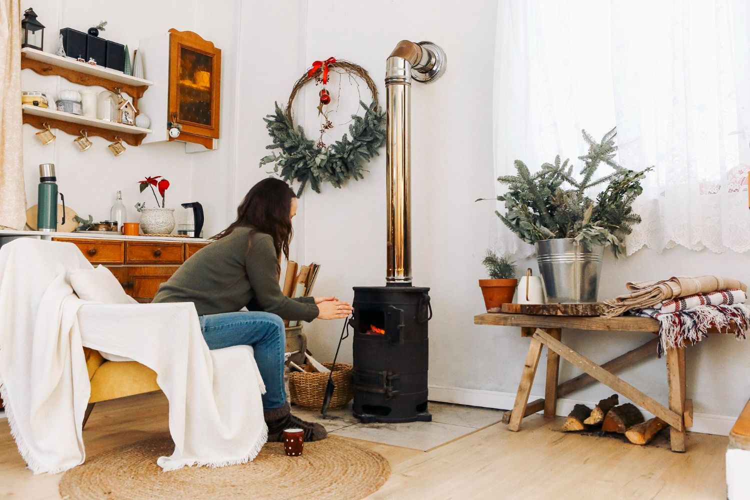 Person tending a wood stove in a cozy, rustic living room with plants and holiday decor