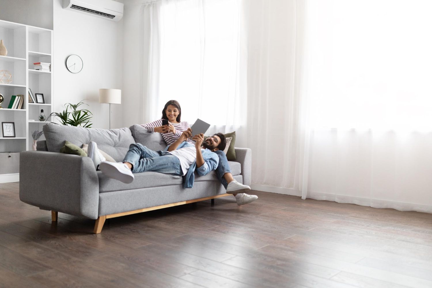 Two people relaxing on a gray sofa in a bright living room, reading together.