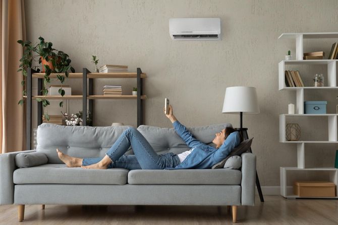 Person lying on a gray sofa in a modern living room with shelves, lamp, and wall-mounted air conditioner.