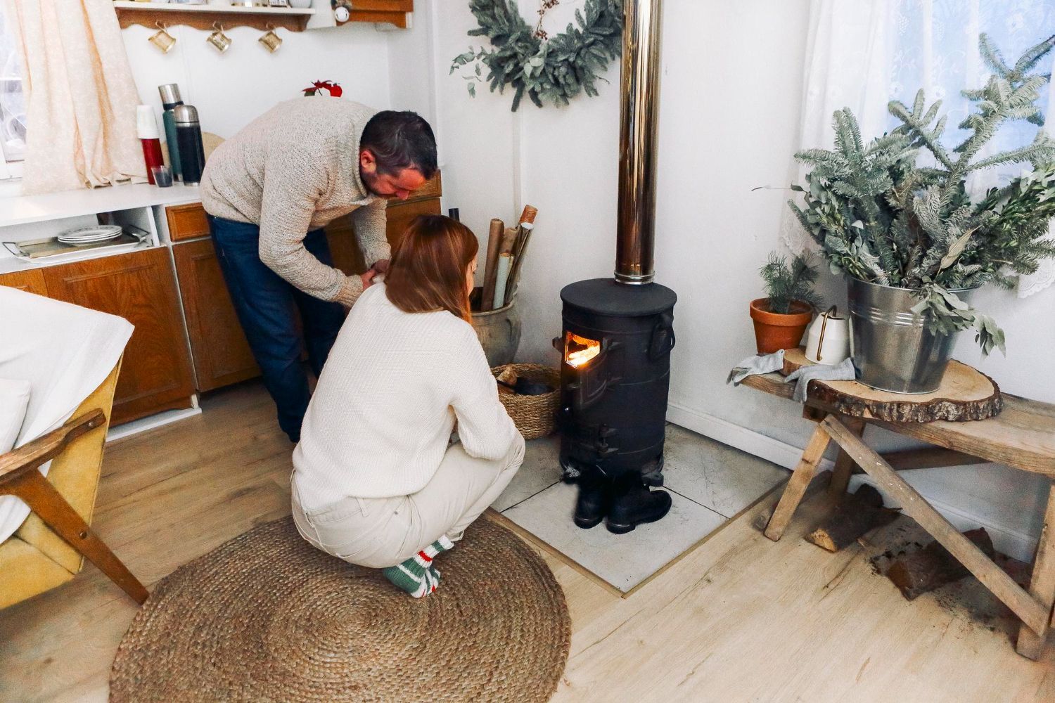 Two people crouch beside a black wood stove in a cozy room with rustic decor and potted plants.