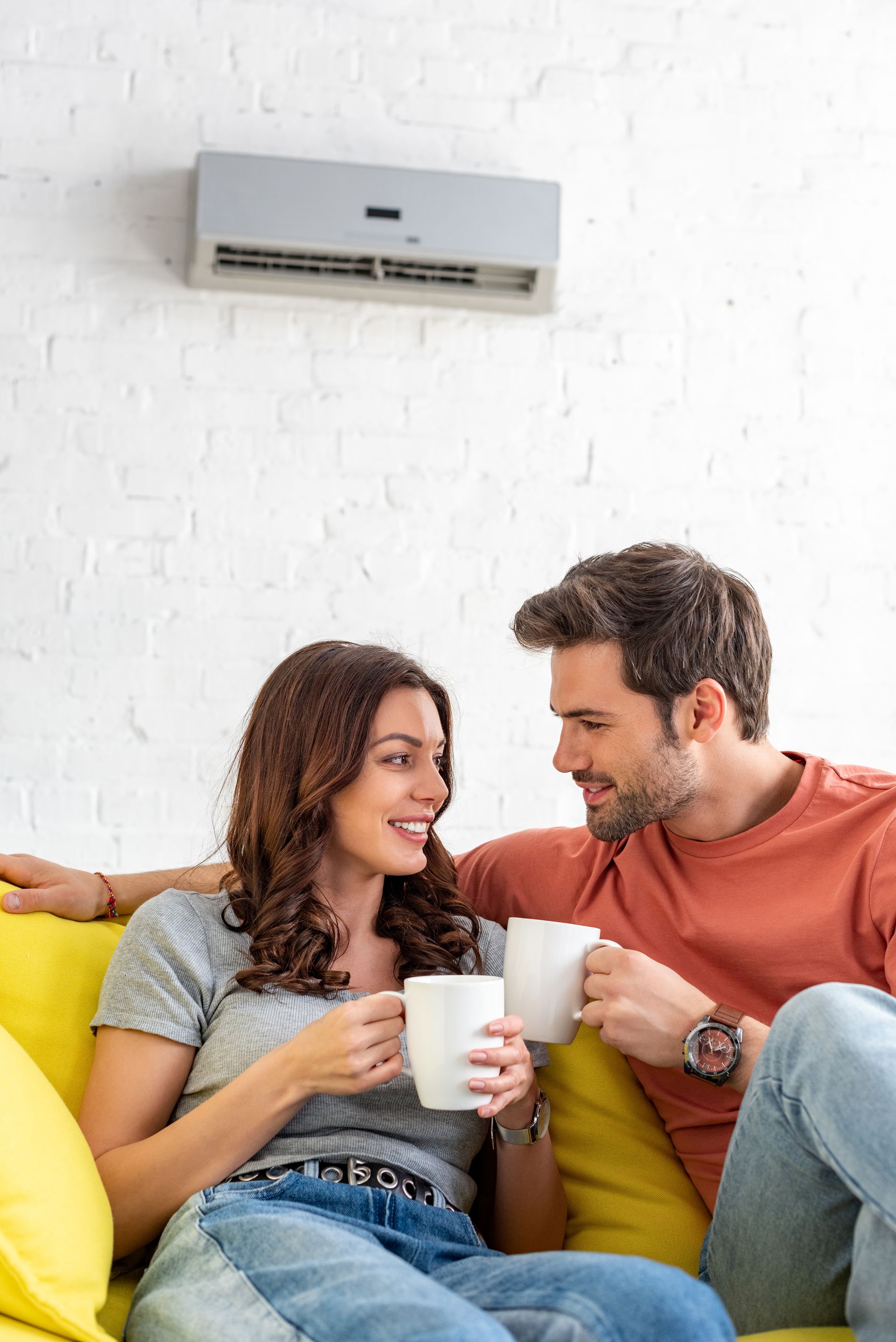Couple relaxing on a yellow couch under a wall-mounted air conditioner, holding mugs and smiling at each other