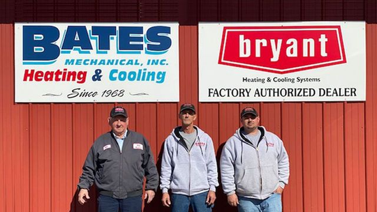 Three workers in jackets stand before Bates Heating & Cooling and Bryant dealer signs on a red building.