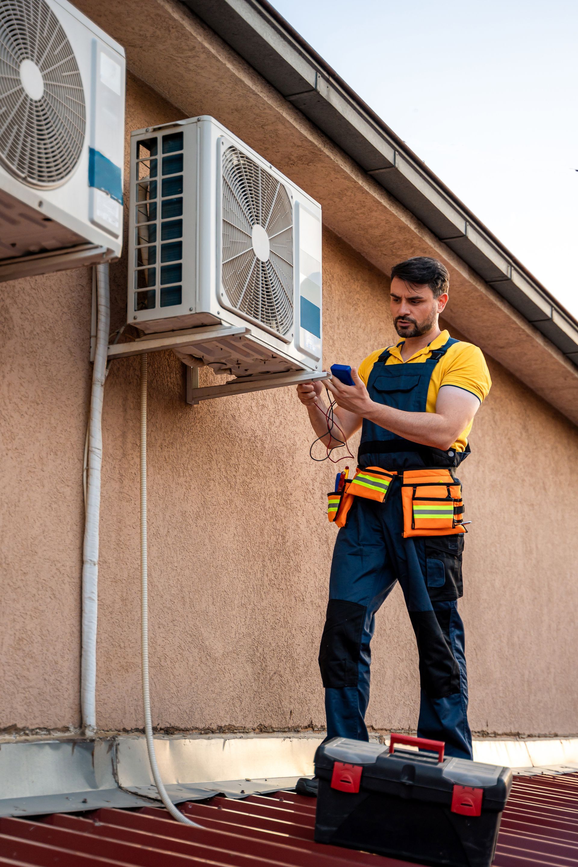Technician inspecting outdoor air conditioner units from a roof ladder with tools on a work belt