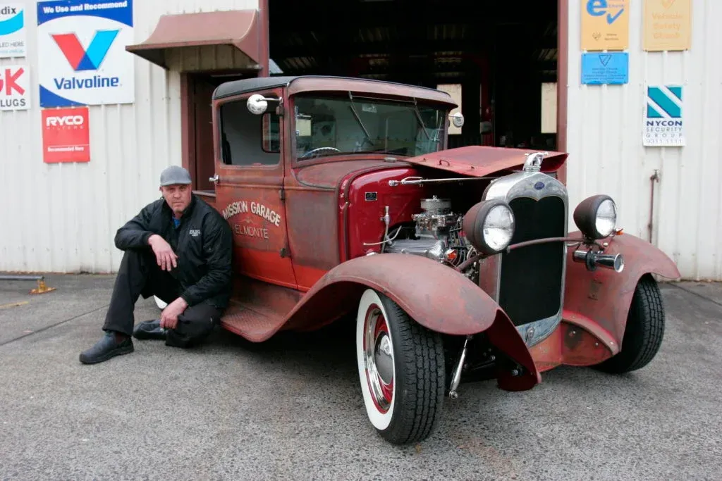 Man sits next to a rusty, vintage pickup truck in front of a garage — Bob Kolln Automotive In Charmhaven, NSW