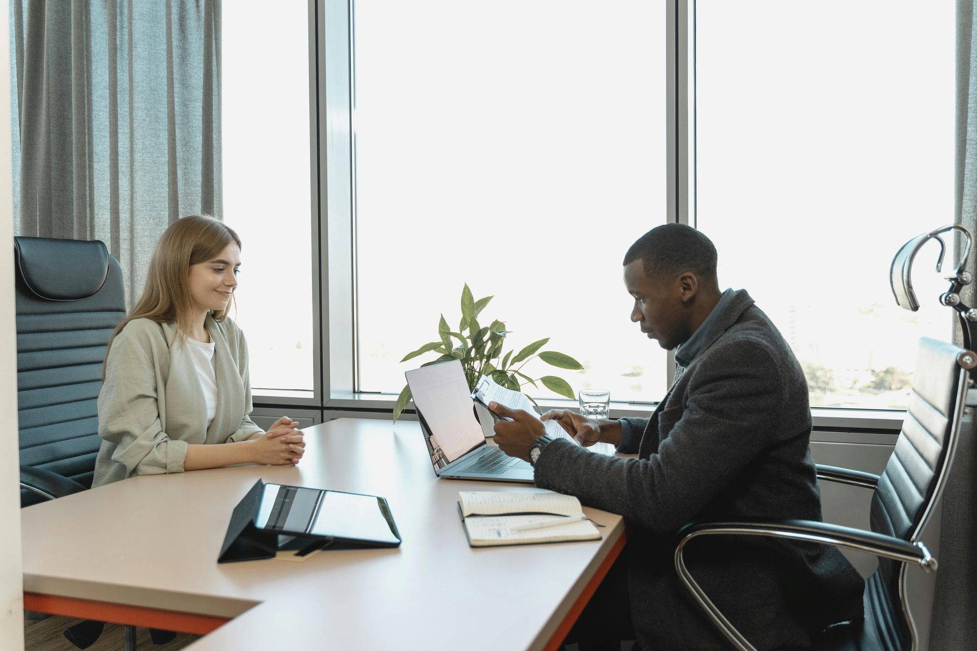 Two people in a bright office meeting across a desk with a laptop, notebook, and potted plant.