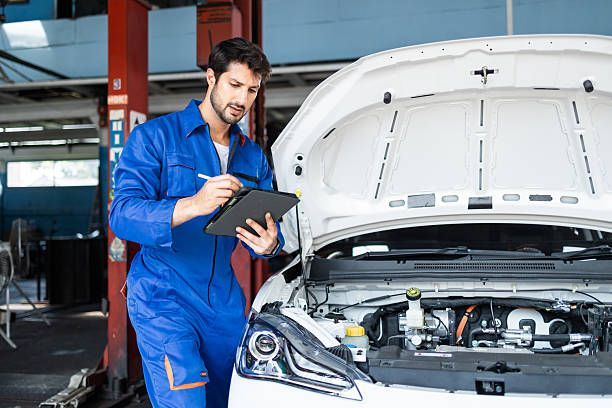 Mechanic in blue coveralls using a tablet beside a car with its hood open in a garage