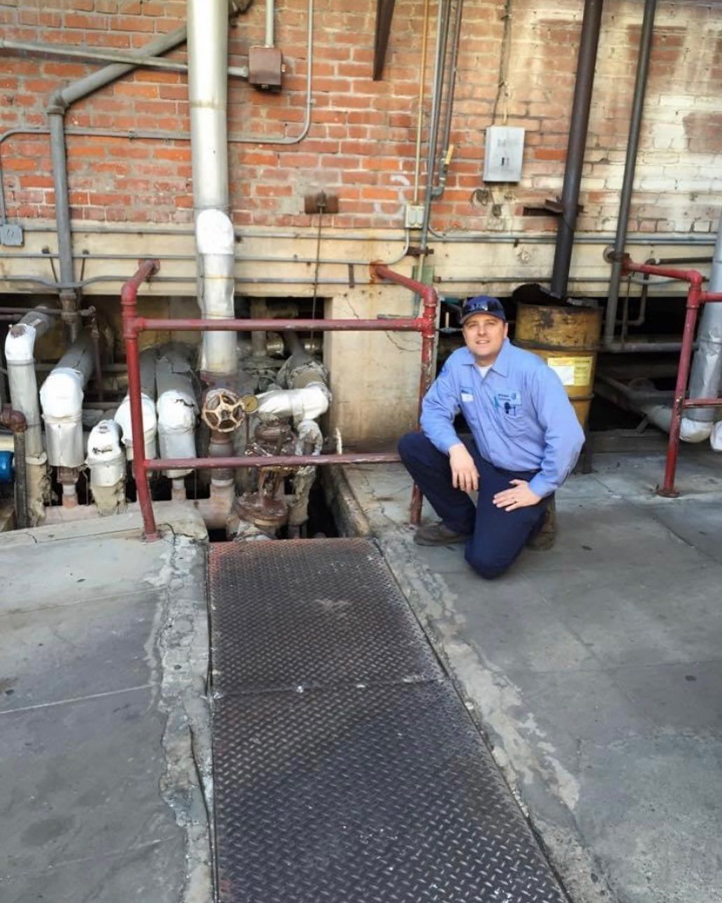 A man in a blue shirt is kneeling down in front of pipes