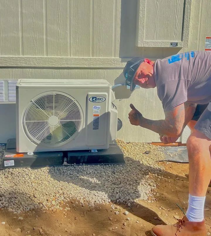 Technician kneels beside an outdoor HVAC unit, giving a thumbs-up in a gravel yard.