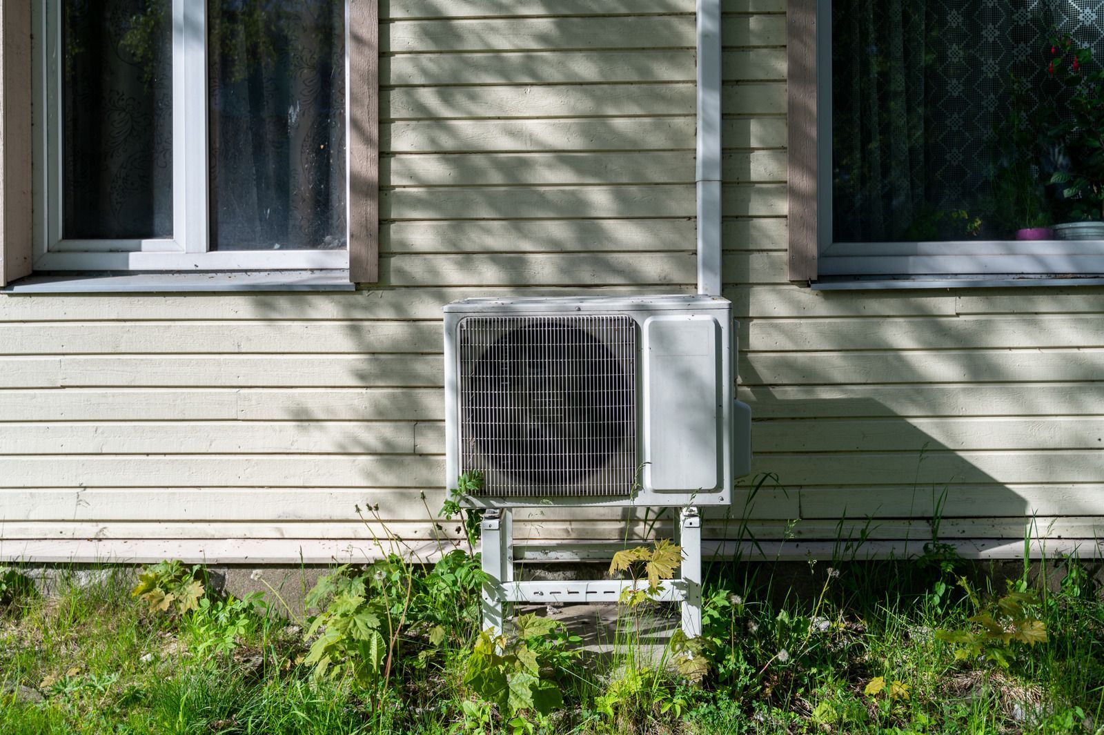 House exterior with two windows and a wall-mounted air conditioner above overgrown grass.