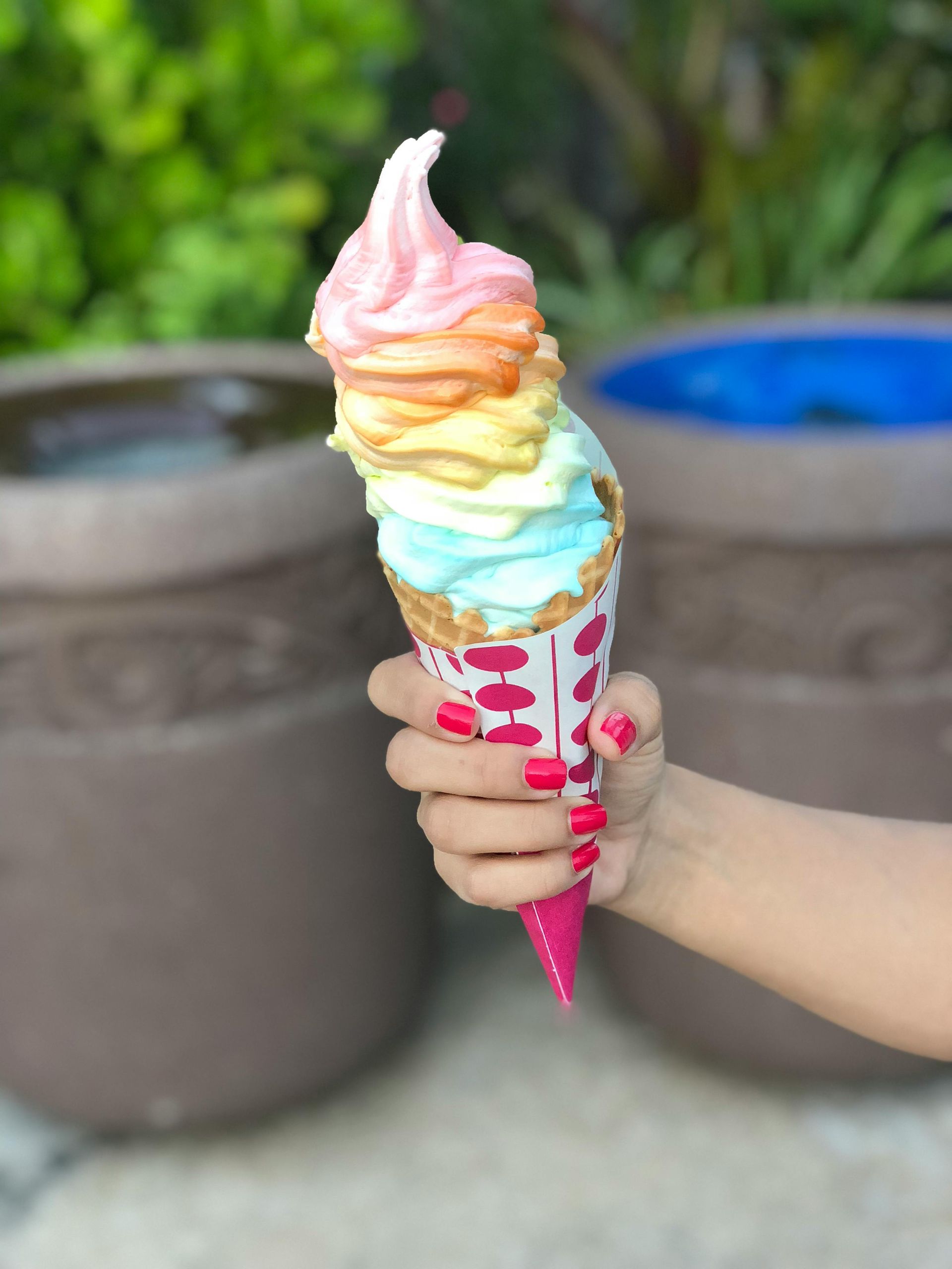 Hand holding a colorful rainbow soft-serve ice cream cone, with pink nails, against a blurred outdoor background.