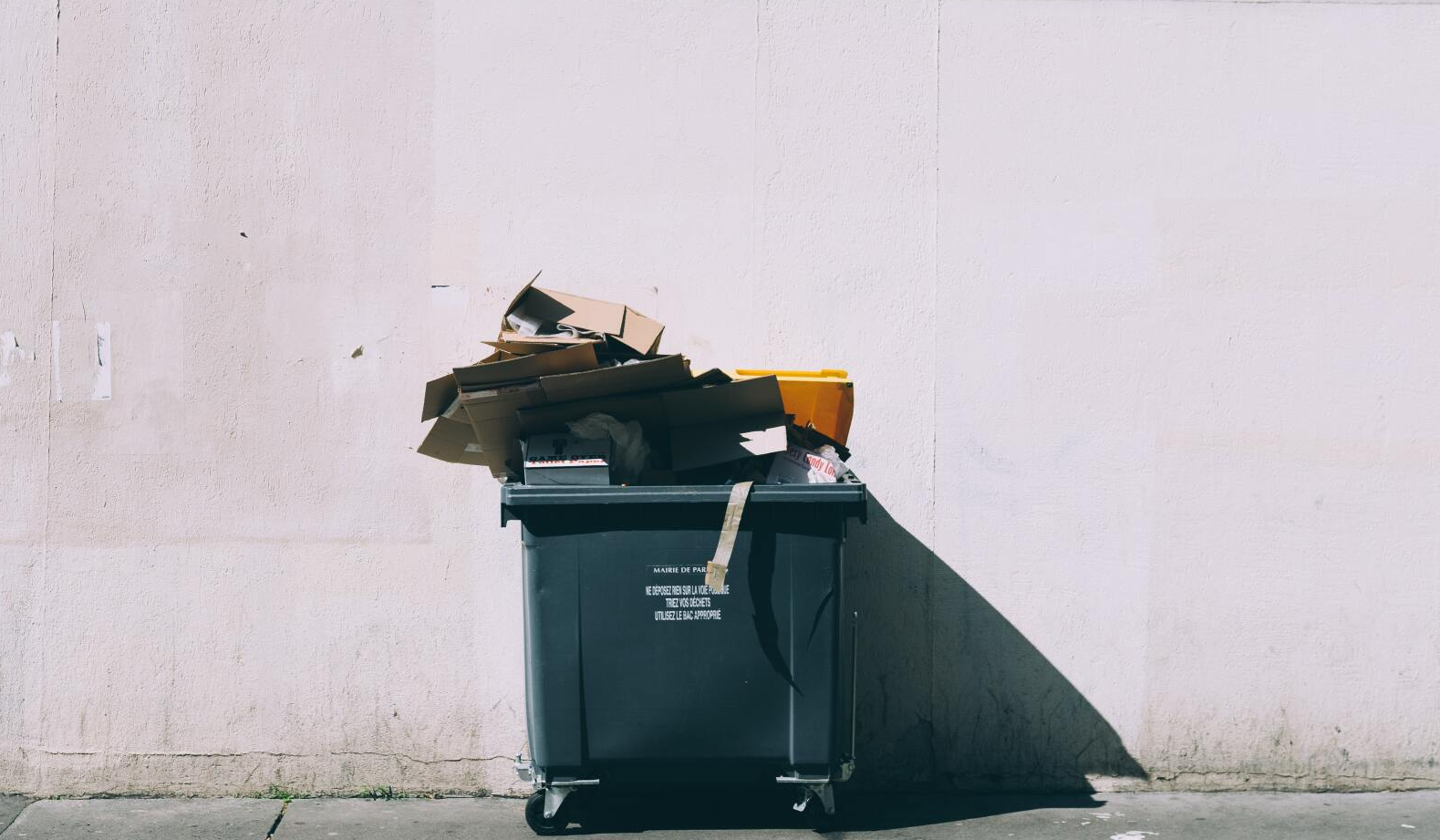 A trash can filled with garbage is sitting on the sidewalk in front of a wall.