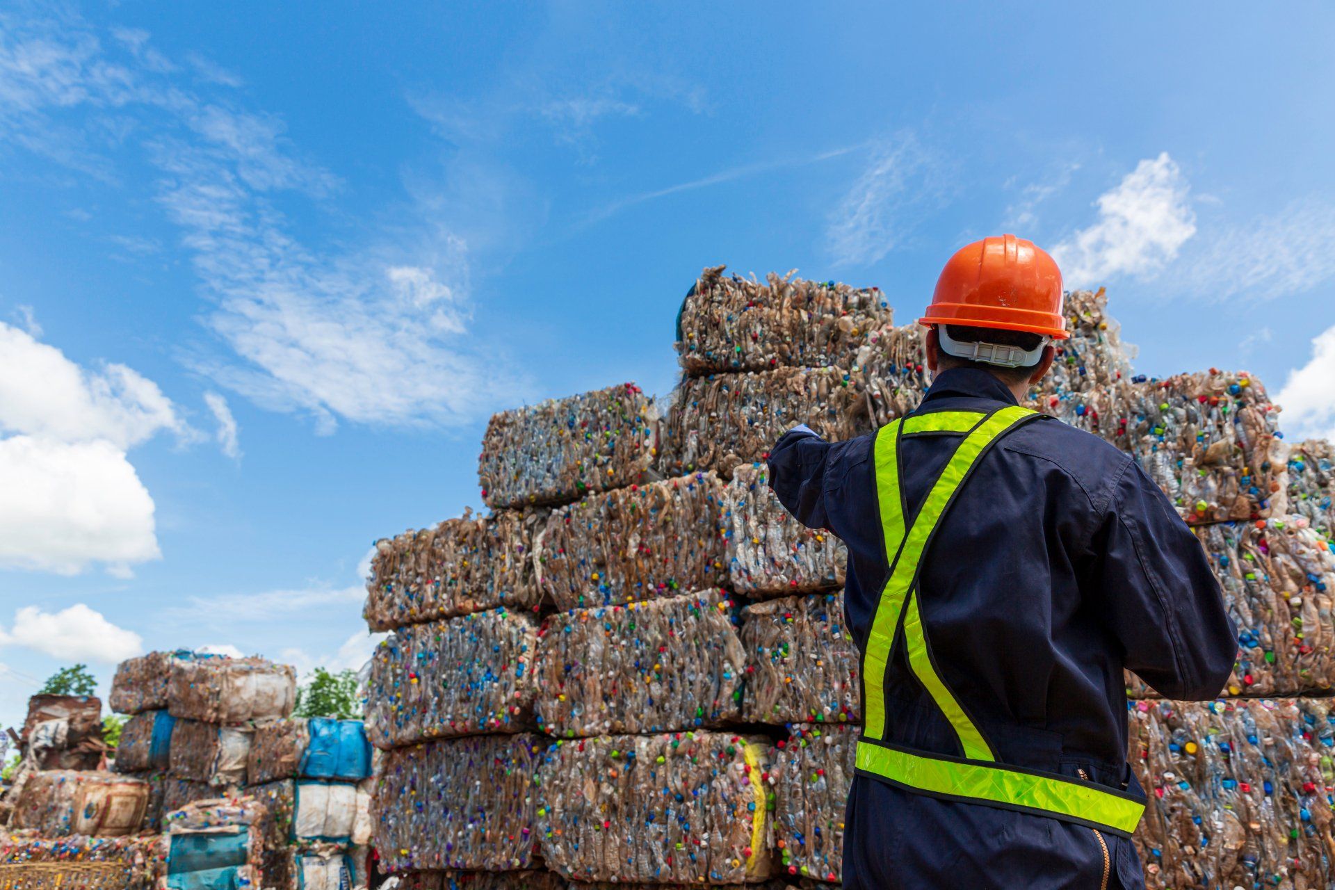 A man wearing a hard hat and safety vest is standing in front of a pile of plastic bottles.