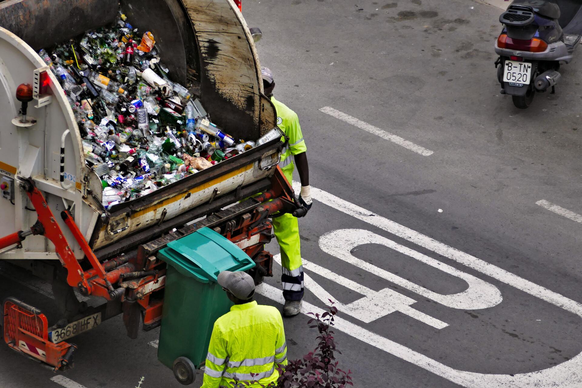 A garbage truck is full of trash and a scooter is driving by