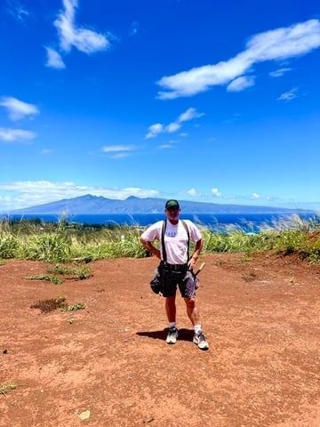 Person standing on a red dirt overlook with mountains and blue sky in the background