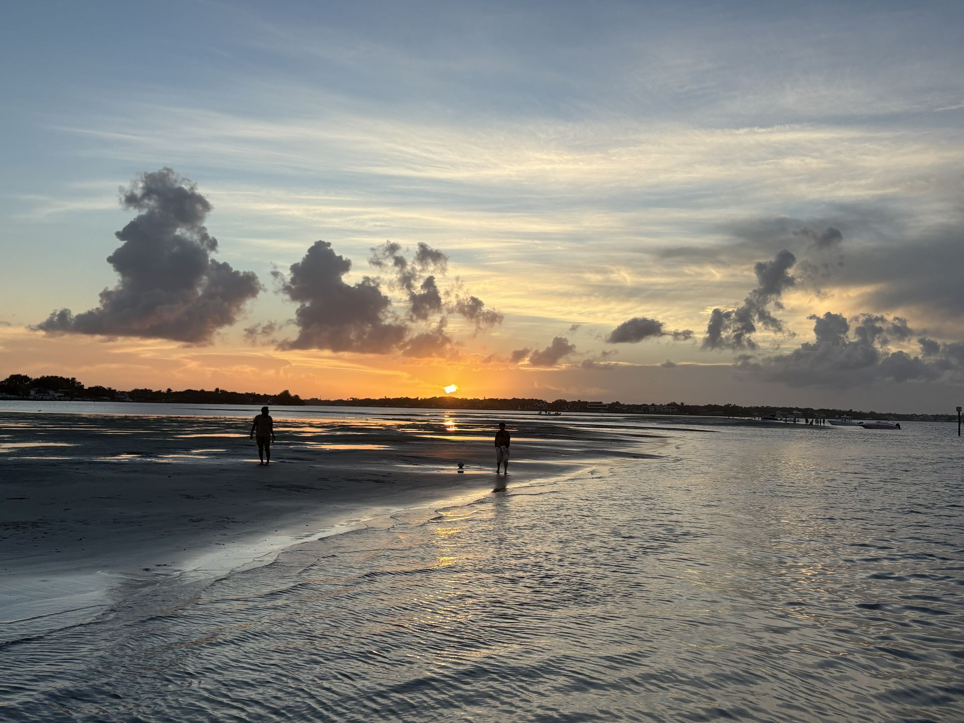 Sunset beach with two silhouetted walkers, reflective wet sand, and dramatic clouds over calm water.