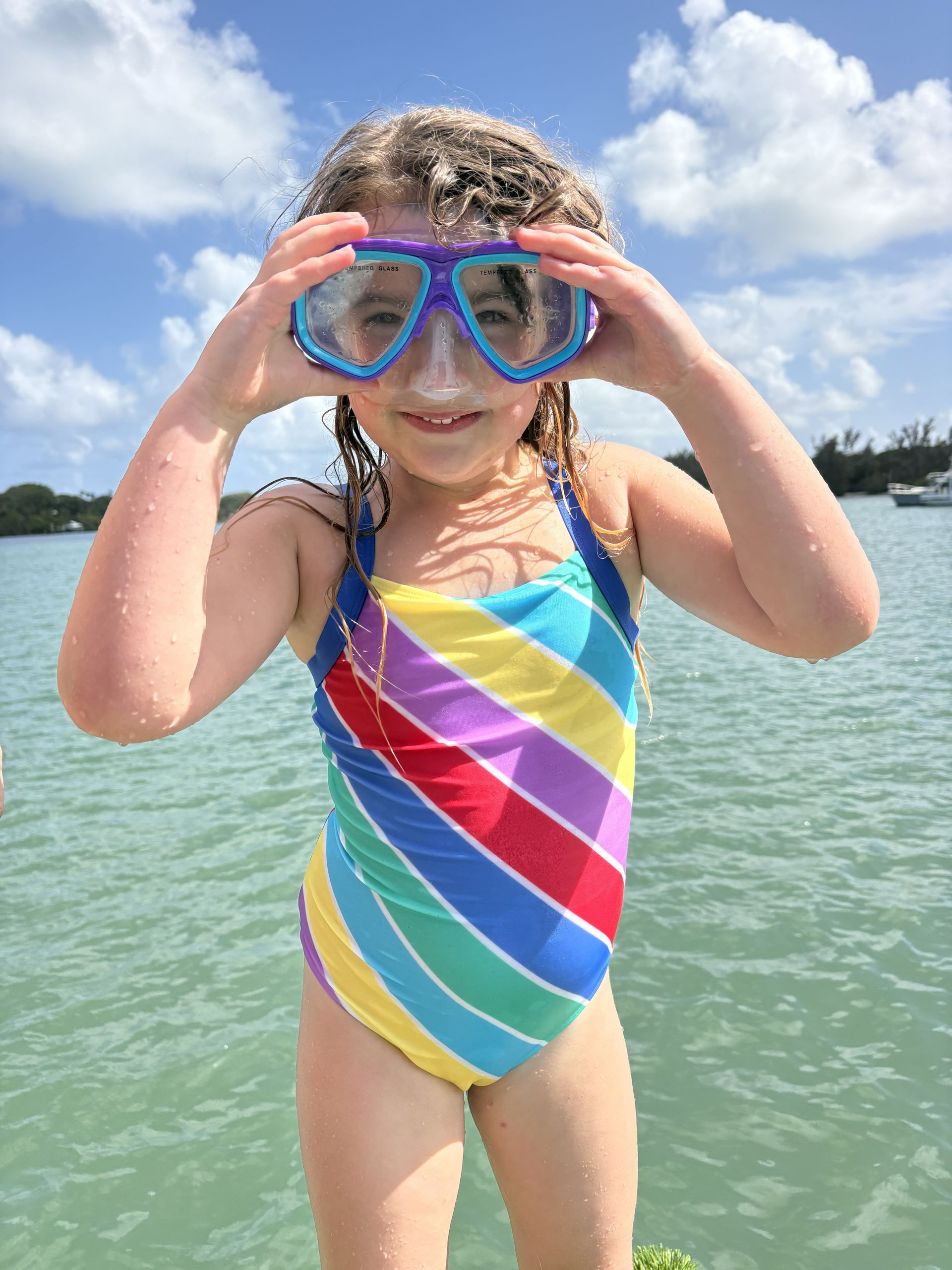 Child in a colorful striped swimsuit adjusts blue goggles while standing in shallow water.