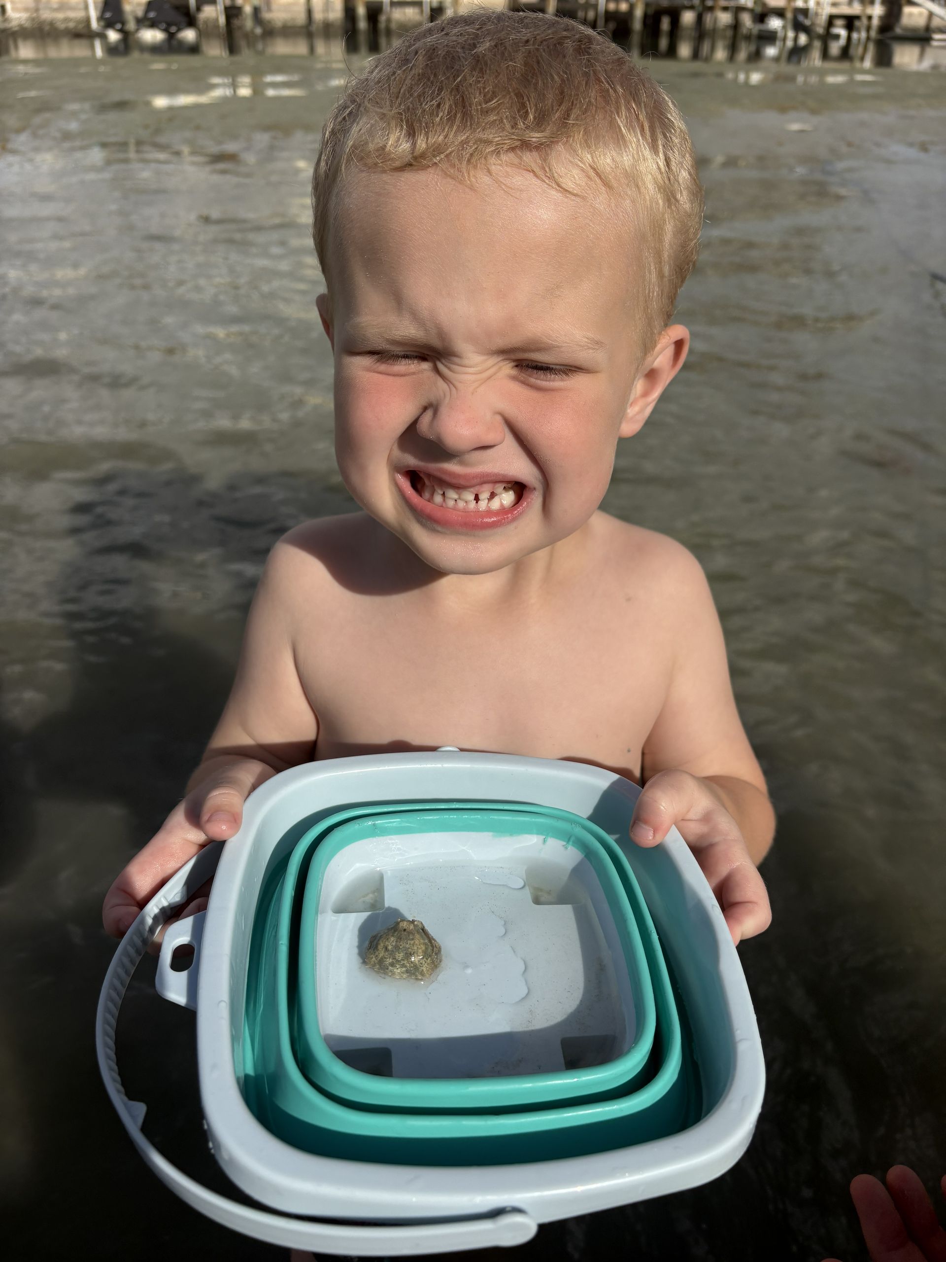 Toddler at the beach holding stacked containers, squinting with a strained expression in shallow water.