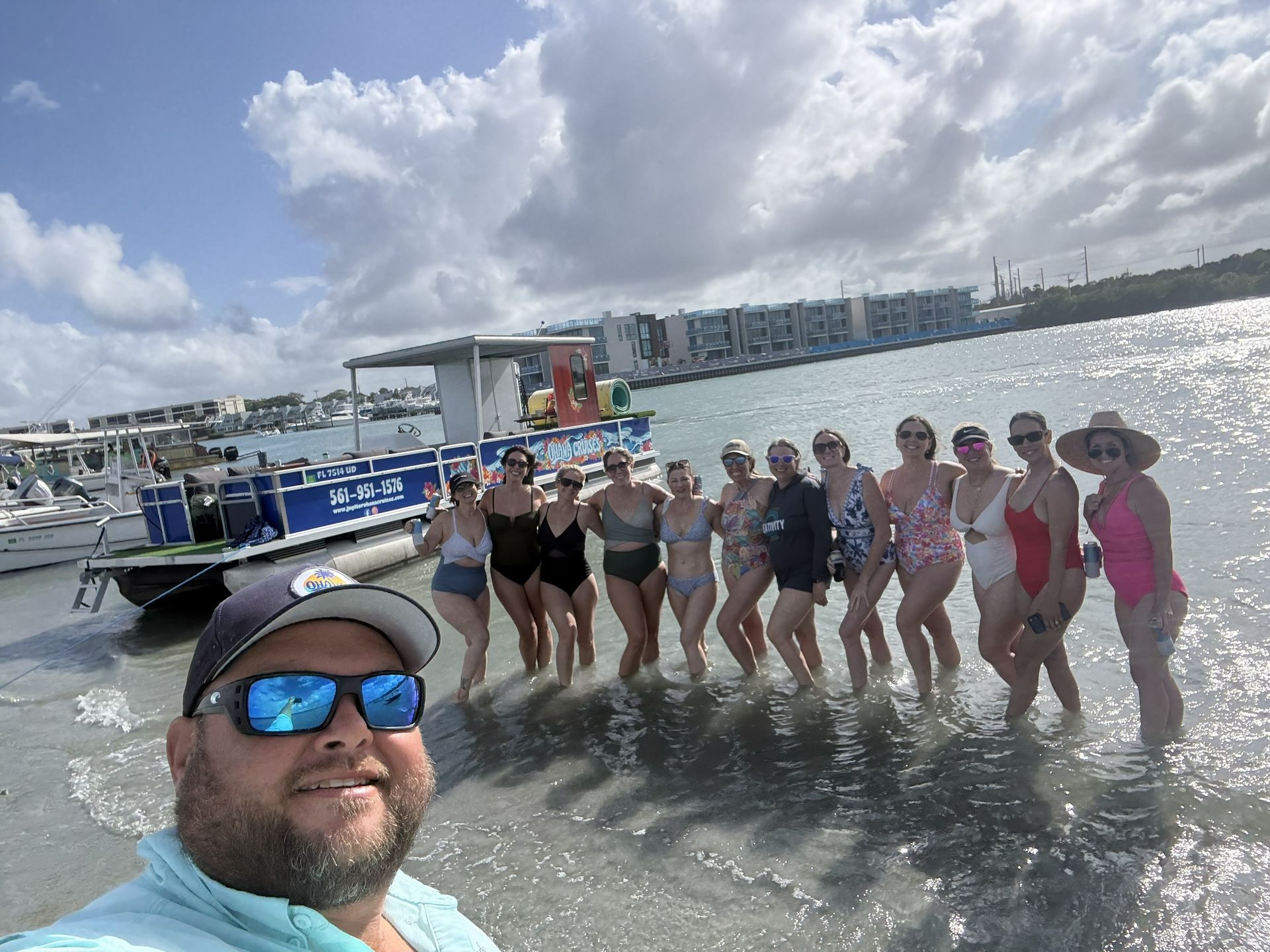Group posing in shallow water beside a boat, with a man taking a selfie in the foreground under a cloudy sky