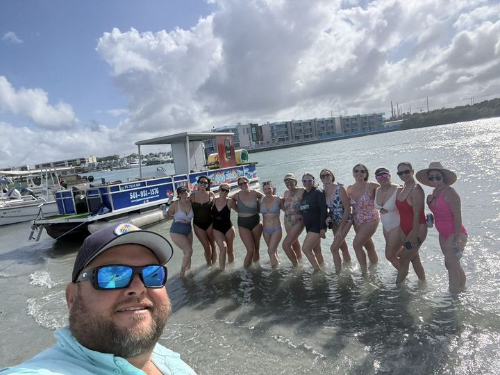 Group posing in shallow water by a docked boat, with a man taking a selfie in the foreground.