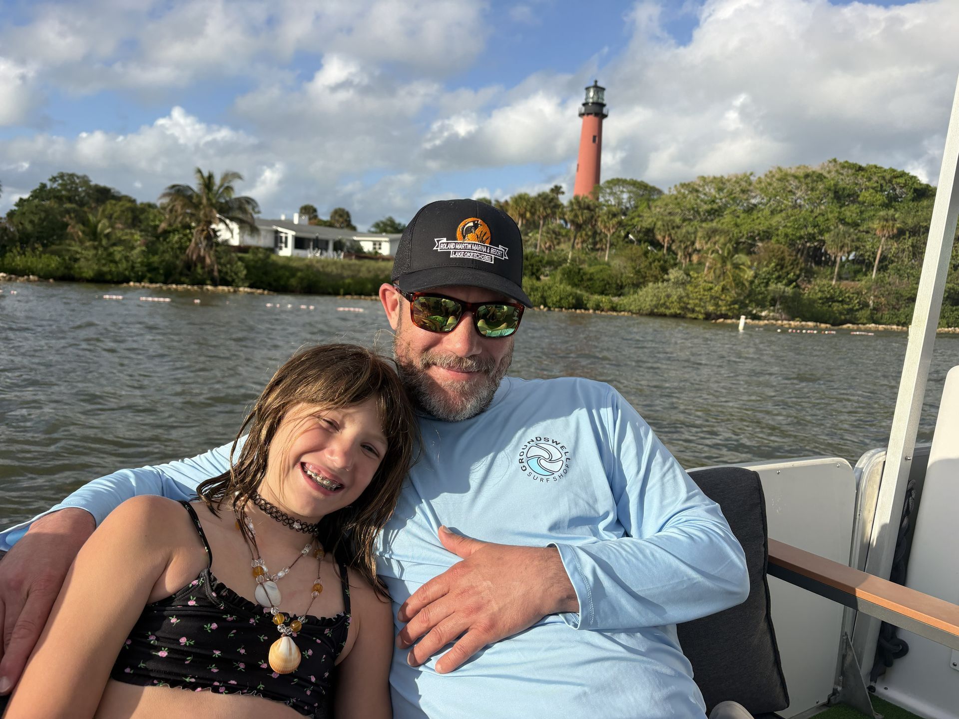 Two people smiling on a boat with a red lighthouse and shoreline in the background.