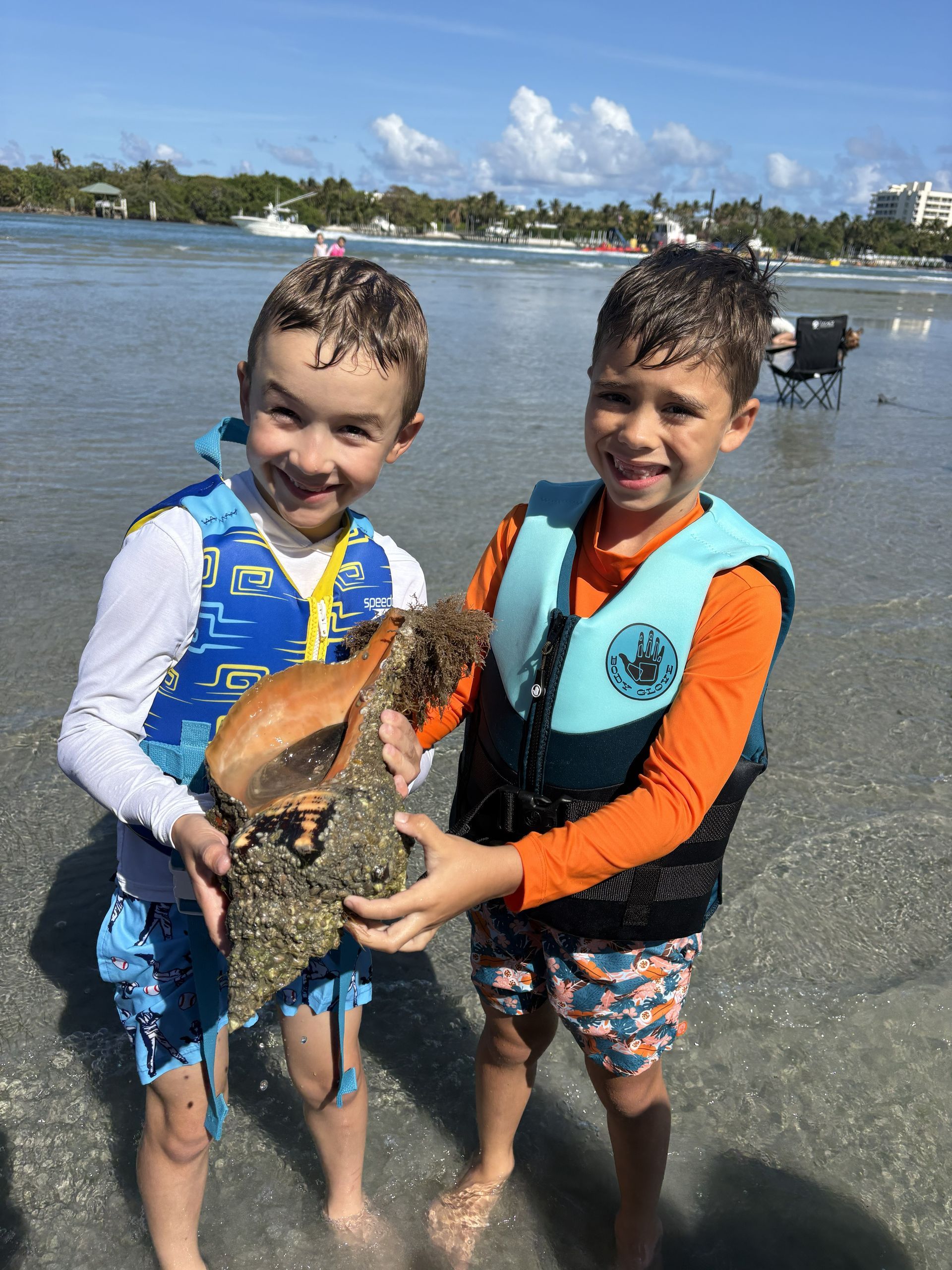 Two children in life vests hold a fish on a beach shoreline.