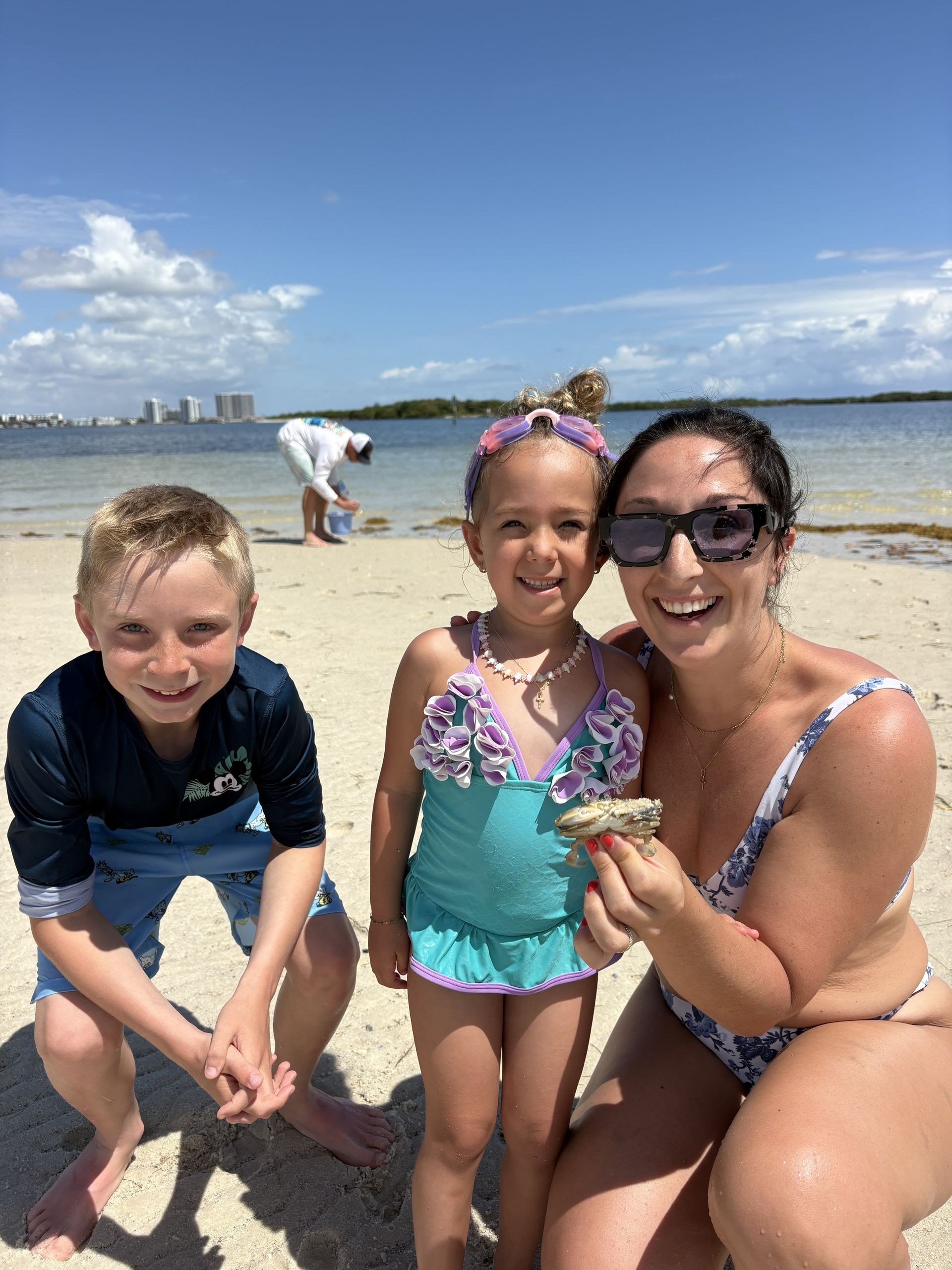 Three people posing on a sunny beach, smiling and crouching by the shoreline.
