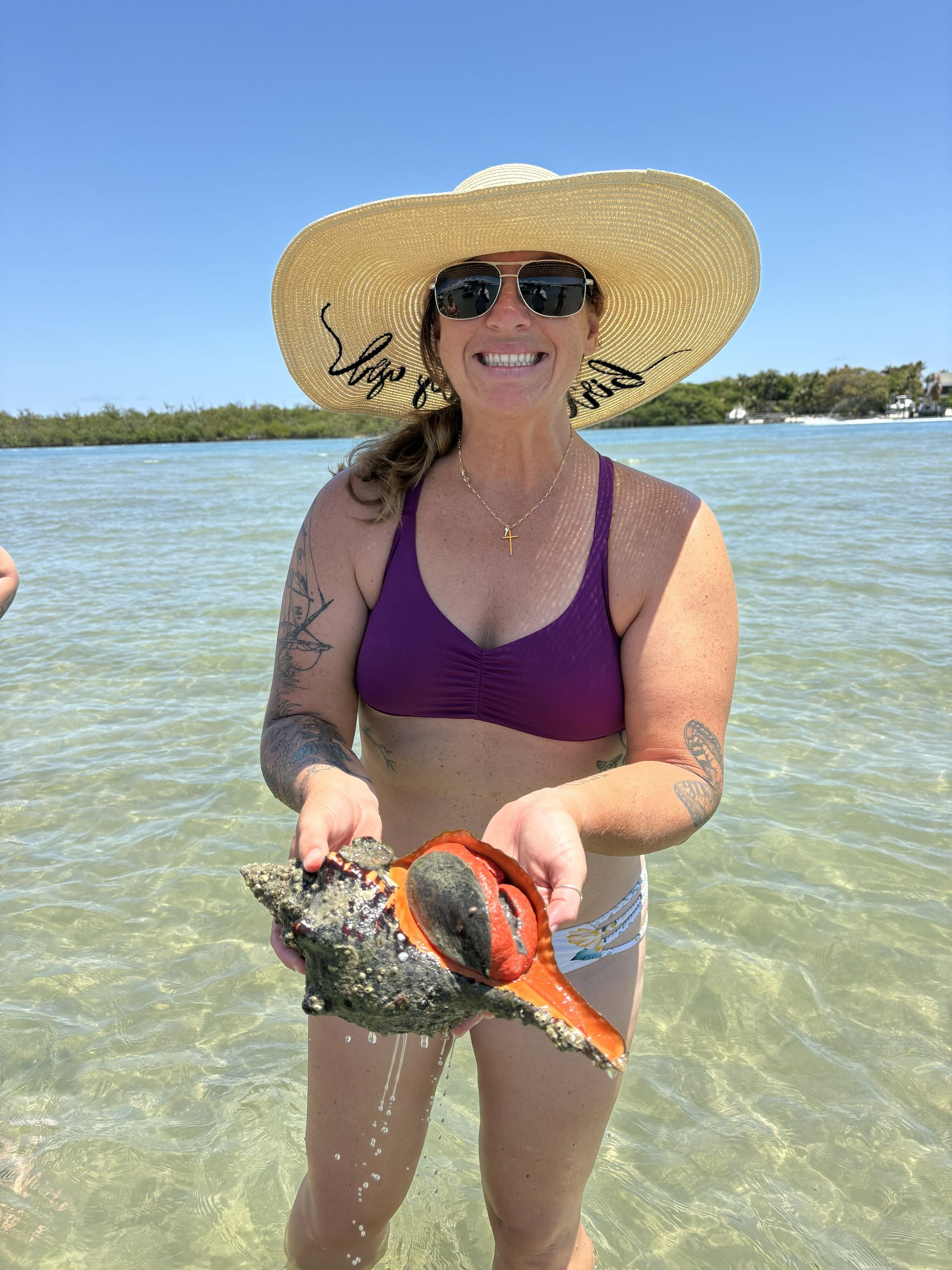 Woman in sunglasses and sunhat holding a large shell while standing in shallow clear water