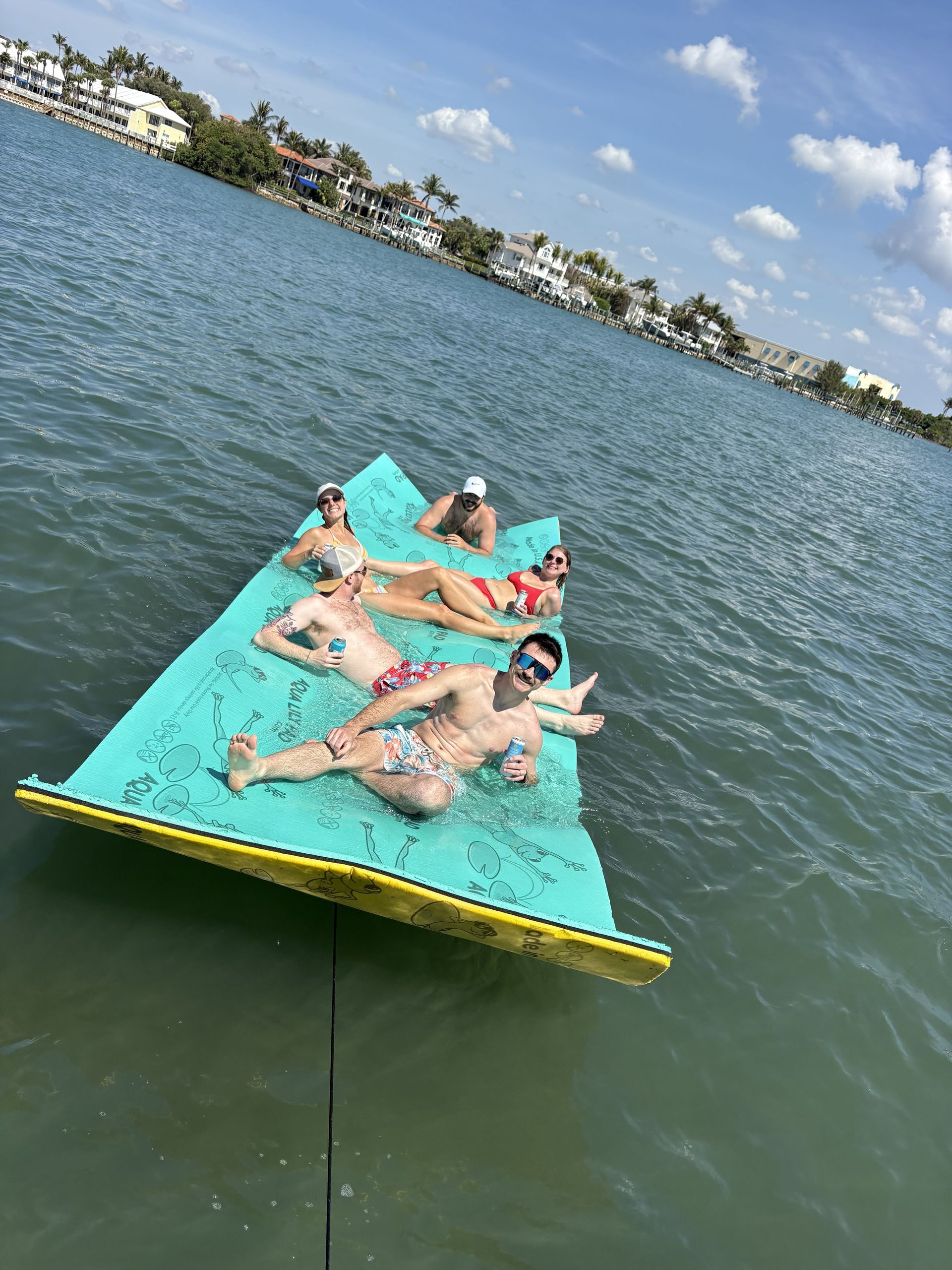 People lounging on a turquoise platform floating on a lake, with shoreline buildings in the background.