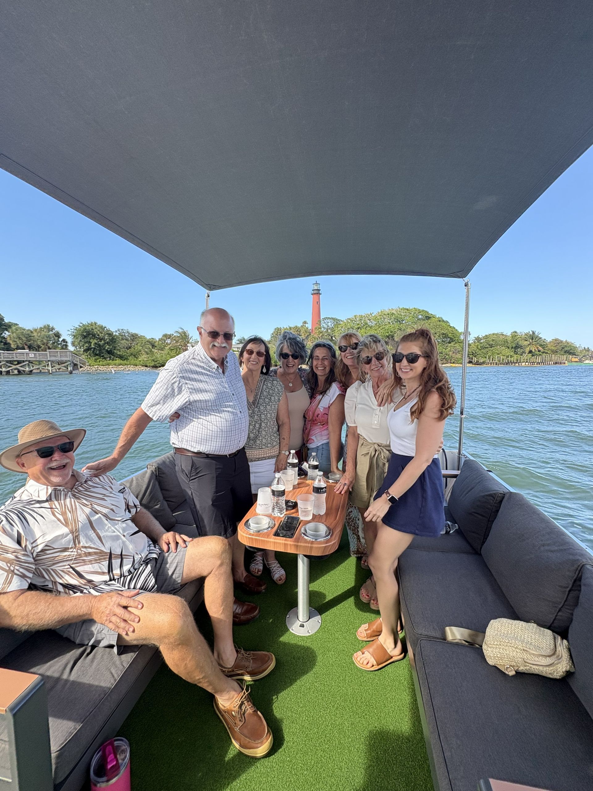 Group gathered on a shaded boat deck with drinks, smiling around a table on a sunny day.